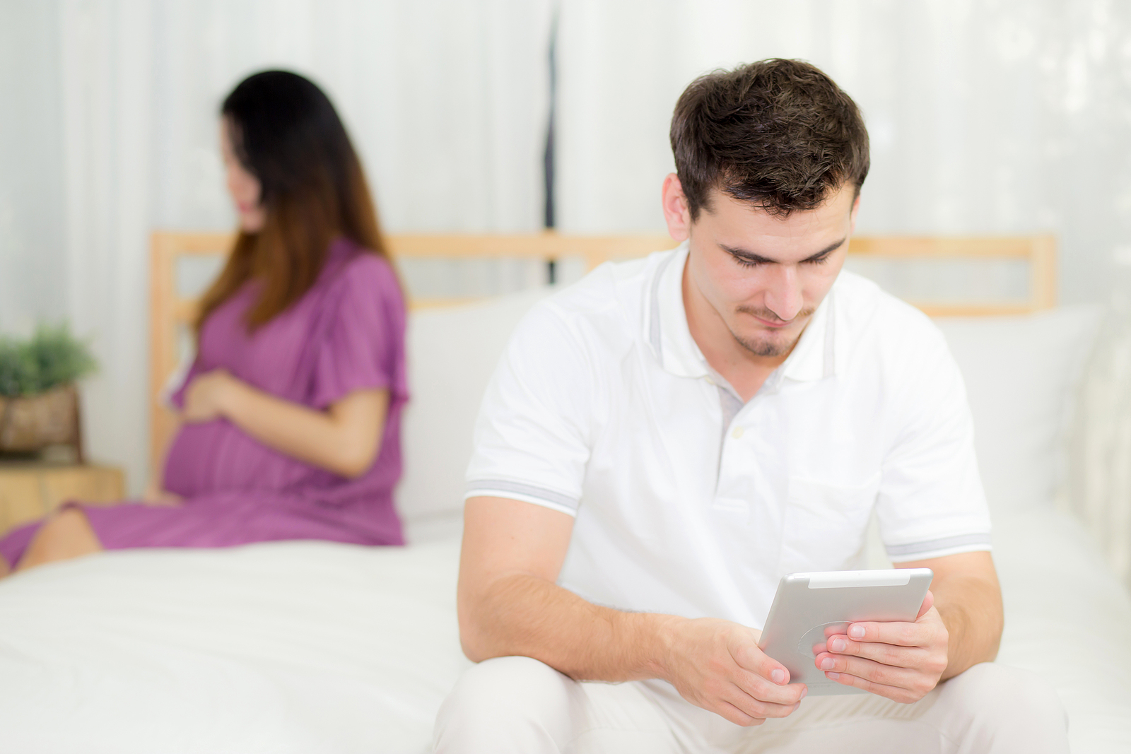 Young couple sitting on a bed, with a pregnant woman in the background and a man using a tablet in the foreground.